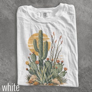 May include: A white short-sleeve t-shirt featuring a graphic print of a desert scene with a tall saguaro cactus, prickly pear cacti, and agave plants in shades of green and orange against a large yellow sun. The inner neck label reads Comfort Colors.