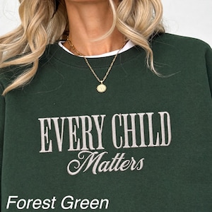 以下が含まれることがあります： 胸元に白い刺繍で「EVERY CHILD Matters」と施されたフォレストグリーンのクルーネックトレーナー。縁に質感のある円形のペンダントと中央に文字があしらわれた、ゴールドトーンのチェーンネックレスを着用しています。