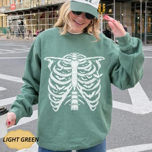 May include: A light green crewneck sweatshirt featuring a large white graphic print of a human rib cage on the front. The rib cage design is composed of numerous small bat silhouettes integrated into the bone structure, with long sleeves.