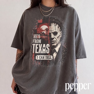 May include: A charcoal grey short-sleeve t-shirt featuring a graphic print of a masked character. The design includes a red and black landscape, the text "HE'S FROM TEXAS I CAN TELL," and a detailed portrait of a man wearing a stitched mask.