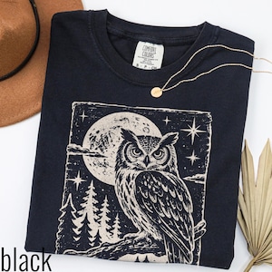 May include: A black short-sleeve t-shirt featuring a cream-coloured graphic print of an owl perched on a branch in front of a large moon, stars, and pine trees. The inner neck label reads Comfort Colors, 100% Cotton, Made in Honduras, size S.