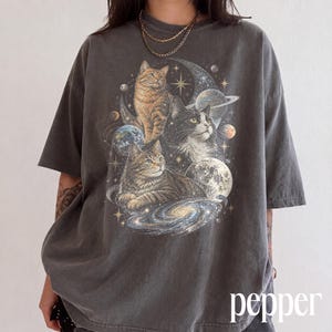 Könnte beinhalten: Ein anthrazitfarbenes Kurzarm-T-Shirt mit einem Grafikdruck von drei Katzen, umgeben von himmlischen Elementen wie Planeten, Sternen, einer Mondsichel und einer Spiralgalaxie, die mittig auf der Brust des Kleidungsstücks platziert sind.