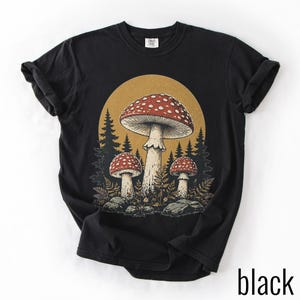 Può includere: Una t-shirt nera a maniche corte con una stampa grafica di tre funghi rossi a pois bianchi in un ambiente boschivo. Il design presenta un grande fungo centrale affiancato da due più piccoli su uno sfondo circolare color oro.