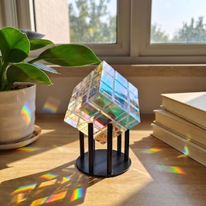 May include: A translucent, iridescent cube with a grid pattern, resembling a puzzle, resting on a black circular stand with four vertical supports. The cube reflects light, creating rainbow-colored prisms on the surface beneath it.