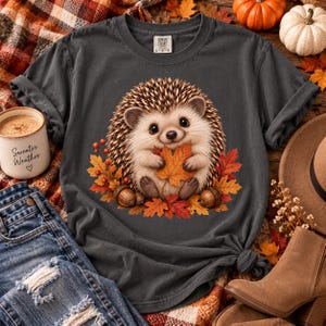 May include: A charcoal gray short-sleeve t-shirt featuring a graphic illustration of a cute hedgehog holding an orange autumn leaf, surrounded by red and orange fall foliage and small acorns. The shirt has a crew neckline and a Comfort Colors label.