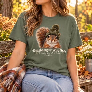 Könnte beinhalten: Ein olivgrünes T-Shirt mit einer Grafik eines braunen Eichhörnchens, das eine dunkelgrüne Strickmütze trägt. Unter dem Eichhörnchen steht der Text "Rehabbing the Wild Ones" gefolgt von "one tiny life at a time" mit einem kleinen Herzsymbol.