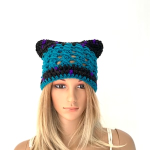 May include: A teal blue crocheted beanie with black and purple accents. The beanie has two ear flaps that are folded down.