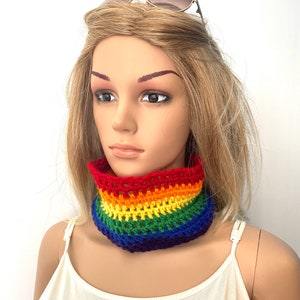 Può includere: Un scaldacollo a uncinetto arcobaleno con strisce rosse, arancioni, gialle, verdi, blu e viola. Lo scaldacollo è indossato da un manichino con i capelli biondi.