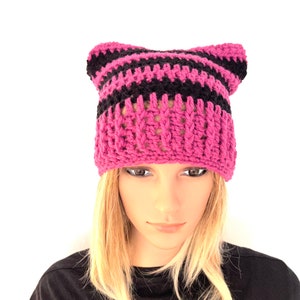 May include: A pink and black striped crochet hat with cat ears. The hat is made of a chunky yarn and has a ribbed brim. The cat ears are pointed and stand up from the top of the hat.