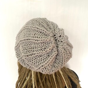 Op de afbeelding: Een grijze gebreide beanie met een slouchy pasvorm. De beanie is gemaakt met een eenvoudig steekpatroon en heeft een ontspannen, casual look.