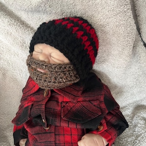 Op de afbeelding: Een baby draagt ​​een zwarte en rode gehaakte muts met een bruine gehaakte baard eraan vast. De baby draagt ​​een rood en zwart geruit overhemd.