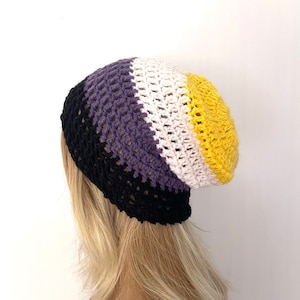 Puede incluir: Un gorro de punto de ganchillo con rayas negras, moradas, blancas y amarillas. El gorro es holgado y tiene un ajuste holgado.