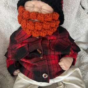 Op de afbeelding: Een baby die een rood-zwart geruit overhemd draagt, een zwart-rode gehaakte muts en een oranje gehaakte baard.