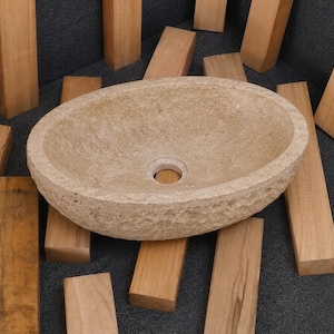Puede incluir: Un lavabo ovalado hecho de piedra natural de color beige claro. El exterior presenta un acabado rugoso y texturizado, mientras que la cubeta interior es lisa con un orificio de drenaje circular central. La piedra muestra variaciones naturales.