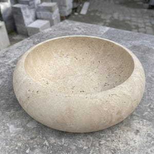 Może przedstawiać: Okrągła, beżowa kamienna umywalka nablatowa o porowatej, naturalnej fakturze. Misa posiada gładką, zaokrągloną krawędź oraz głębokie, wklęsłe wnętrze, ukazujące organiczne wzory i jasnobrązowy odcień charakterystyczny dla naturalnego trawertynu.