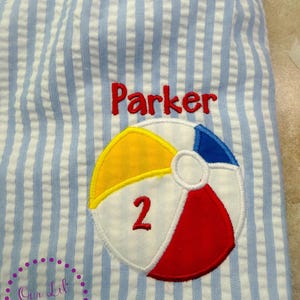 Puede incluir: Tela de seersucker azul claro y blanca con una pelota de playa bordada con paneles amarillos, azules y rojos. El n&uacute;mero 2 est&aacute; cosido en rojo sobre la pelota de playa, y el nombre Parker est&aacute; bordado en rojo sobre el dise&ntilde;o de la pelota.