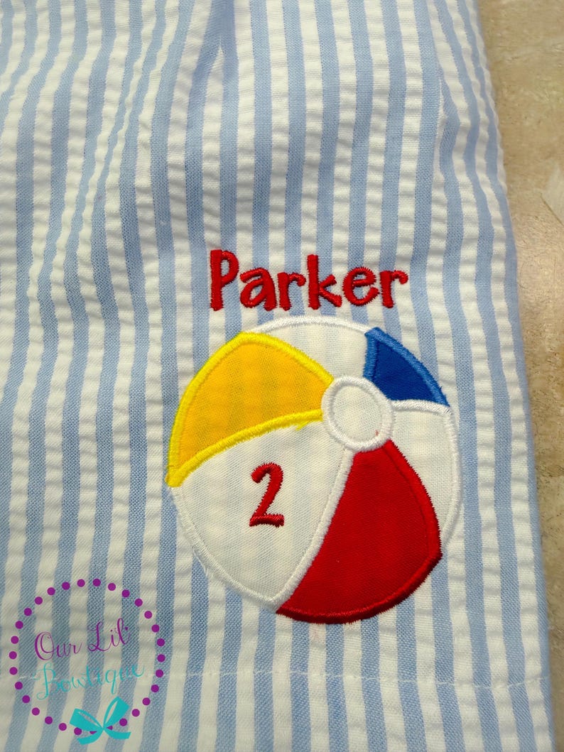 Puede incluir: Tela de seersucker azul claro y blanca con una pelota de playa bordada con paneles amarillos, azules y rojos. El n&uacute;mero 2 est&aacute; cosido en rojo sobre la pelota de playa, y el nombre Parker est&aacute; bordado en rojo sobre el dise&ntilde;o de la pelota.