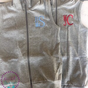 May include: Two gray zip-up vests with embroidered initials. One vest has blue initials "BSM", the other has red initials "MJC". Both vests have a checkered pattern lining the collar. The vests are displayed on a light wood surface.