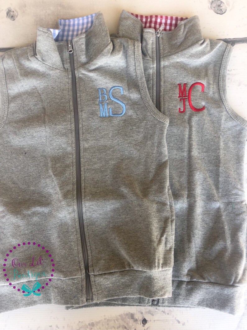 May include: Two gray zip-up vests with embroidered initials. One vest has blue initials "BSM", the other has red initials "MJC". Both vests have a checkered pattern lining the collar. The vests are displayed on a light wood surface.
