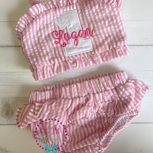 May include: A pink and white striped outfit for a baby's first birthday. The top has a white number one applique with the name "Logan" embroidered in pink. The outfit includes ruffled bloomers.