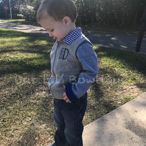 May include: A gray zip-up vest with blue sleeves and a blue and white checkered collar. The vest has the embroidered initials "GD" and is worn with dark blue jeans and navy sneakers.
