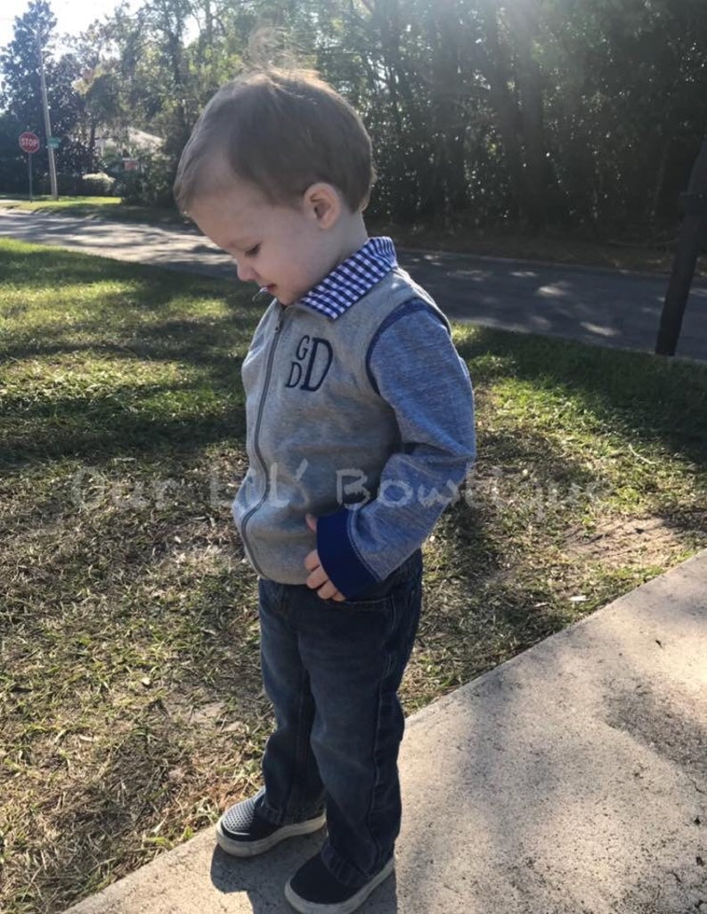 May include: A gray zip-up vest with blue sleeves and a blue and white checkered collar. The vest has the embroidered initials "GD" and is worn with dark blue jeans and navy sneakers.
