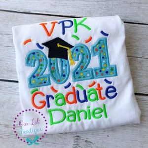 May include: White t-shirt with a blue, green, orange, and yellow embroidered design that says "VPK 2021 Graduate Daniel". A black graduation cap with a yellow tassel is above the year 2021.