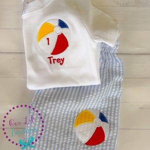 Puede incluir: Una camiseta blanca de manga corta con una pelota de playa bordada que incluye el n&uacute;mero 1 y el nombre Trey en texto rojo, junto a unos pantalones cortos de seersucker azul claro y blanco con una pelota de playa bordada a juego en la pierna.