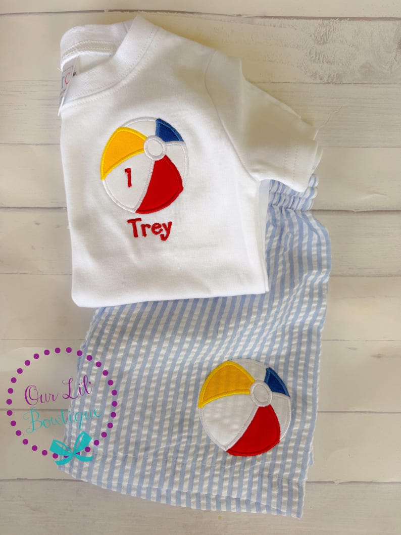 Puede incluir: Una camiseta blanca de manga corta con una pelota de playa bordada que incluye el n&uacute;mero 1 y el nombre Trey en texto rojo, junto a unos pantalones cortos de seersucker azul claro y blanco con una pelota de playa bordada a juego en la pierna.