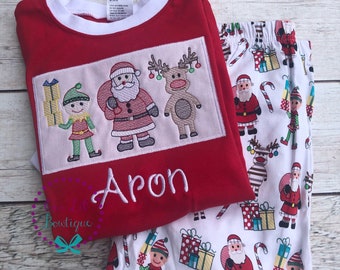May include: Red and white Christmas pyjamas with an embroidered design of Santa, an elf, and a reindeer. The pyjamas have the name "Aron" embroidered on the top.