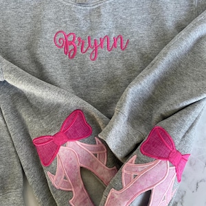 May include: A gray sweatshirt with the name "Brynn" embroidered in pink on the front. The sleeves have pink felt appliques of ballet shoes with bows.