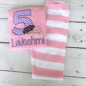 May include: Pink and white striped leggings and a pink shirt with a purple number 5 with white polka dots and a donut graphic. The shirt also has the name "Lakshmi" embroidered on it.