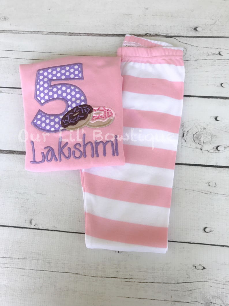May include: Pink and white striped leggings and a pink shirt with a purple number 5 with white polka dots and a donut graphic. The shirt also has the name "Lakshmi" embroidered on it.