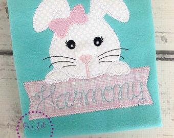 May include: A turquoise blue t-shirt with a white bunny applique with pink polka dots and a pink bow. The bunny is holding a pink banner with the word "Harmony" embroidered on it.