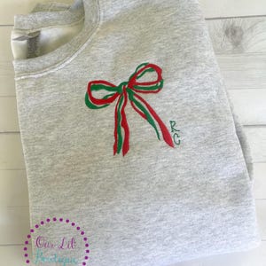 May include: A light gray sweatshirt with a red and green embroidered bow design. The bow is centered on the front and has the initials "RG" embroidered to the right. The sweatshirt is folded and appears to be new.