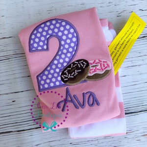 May include: Pink and white striped long-sleeved shirt with a purple polka dot number two and a donut graphic. The shirt says "Ava" in purple embroidery. The shirt has a yellow tag that says "Safety, garment should fit snugly. Garment is not flame resistant. A snug garment is more likely to catch fire."
