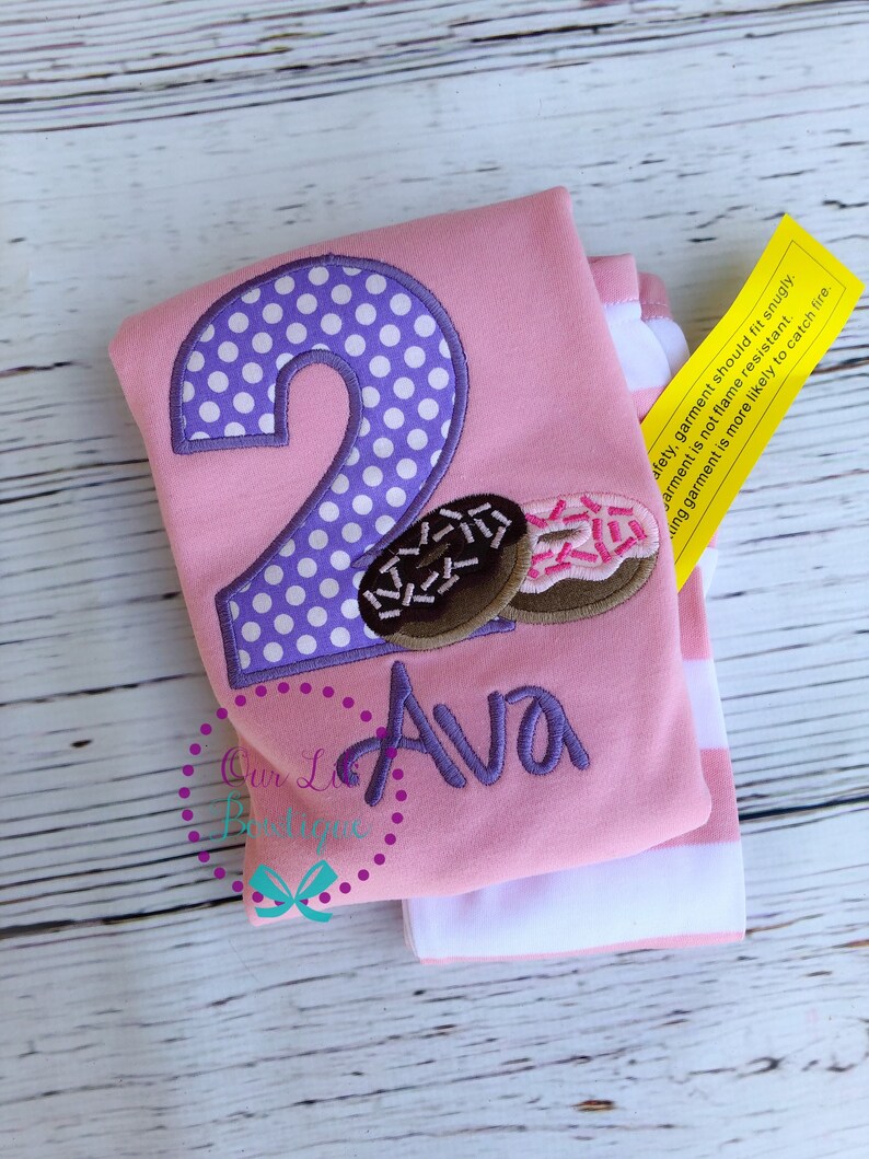 May include: Pink and white striped long-sleeved shirt with a purple polka dot number two and a donut graphic. The shirt says "Ava" in purple embroidery. The shirt has a yellow tag that says "Safety, garment should fit snugly. Garment is not flame resistant. A snug garment is more likely to catch fire."