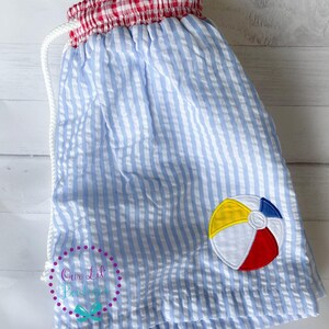 Puede incluir: Pantalones cortos de seersucker azul claro y blanco con cintura el&aacute;stica de cuadros vichy rojos y blancos y cord&oacute;n blanco. Un parche bordado de pelota de playa en amarillo, azul, blanco y rojo est&aacute; en la pierna derecha. Incluye marca de agua.
