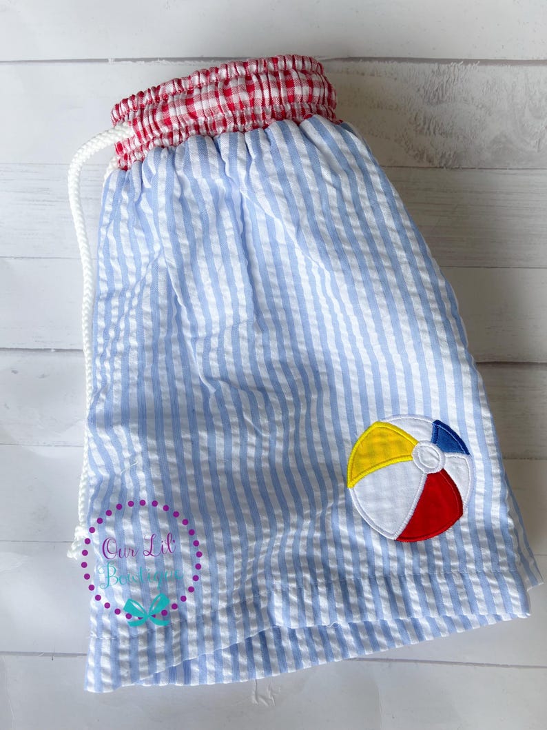 Puede incluir: Pantalones cortos de seersucker azul claro y blanco con cintura el&aacute;stica de cuadros vichy rojos y blancos y cord&oacute;n blanco. Un parche bordado de pelota de playa en amarillo, azul, blanco y rojo est&aacute; en la pierna derecha. Incluye marca de agua.