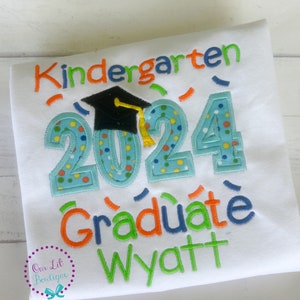 May include: White t-shirt with the text "Kindergarten Graduate 2024" in colorful embroidered letters. A black graduation cap is above the numbers. The name "Wyatt" is embroidered below the text.