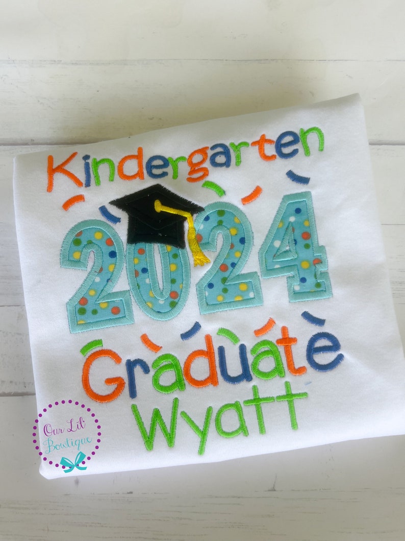 May include: White t-shirt with the text "Kindergarten Graduate 2024" in colorful embroidered letters. A black graduation cap is above the numbers. The name "Wyatt" is embroidered below the text.