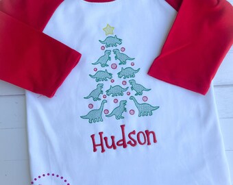 May include: White and red raglan shirt with a Christmas tree design made of green dinosaurs and red ornaments. The name "Hudson" is embroidered in red. The shirt has three-quarter length sleeves and a round neckline.