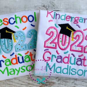 May include: Two white t-shirts with colorful embroidered text and a graduation cap. The left shirt says "Preschool 2022 Graduate Mayson". The right shirt says "Kindergarten 2022 Graduate Madison".