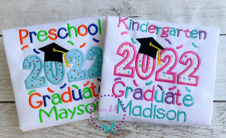 May include: Two white t-shirts with colorful embroidered text and a graduation cap. The left shirt says "Preschool 2022 Graduate Mayson". The right shirt says "Kindergarten 2022 Graduate Madison".