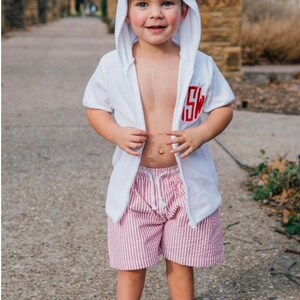 Puede incluir: Una chaqueta blanca de manga corta con capucha y cremallera, con un monograma rojo bordado en el pecho, combinada con pantalones cortos de seersucker a rayas rojas y blancas con cord&oacute;n blanco. El conjunto lo lleva un ni&ntilde;o peque&ntilde;o.