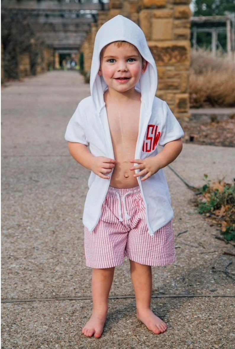 Puede incluir: Una chaqueta blanca de manga corta con capucha y cremallera, con un monograma rojo bordado en el pecho, combinada con pantalones cortos de seersucker a rayas rojas y blancas con cord&oacute;n blanco. El conjunto lo lleva un ni&ntilde;o peque&ntilde;o.