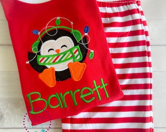 May include: Red pajama set with a white-trimmed top featuring a penguin wearing a scarf and Christmas lights. The penguin has orange feet and a green and white scarf. The name "Barrett" is embroidered in green. The pants are red and white striped.