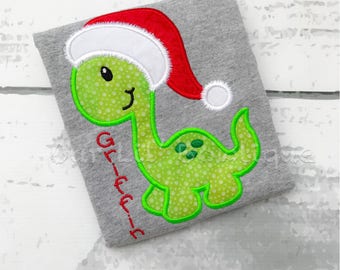May include: A gray t-shirt featuring a green dinosaur wearing a red and white Santa hat. The dinosaur has a polka-dot pattern and is outlined in green. The word "Glee" is written vertically in red.