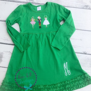 May include: Green long-sleeved dress with a ruffled hem. The dress features an embroidered design of a ballerina, a nutcracker, and a mouse. The dress is monogrammed with the letter "M".