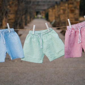 Puede incluir: Tres pares de pantalones cortos de seersucker colgados en un tendedero, cada uno con una cintura de cord&oacute;n blanco. Los pantalones son de color azul claro, verde menta y rosa, con un patr&oacute;n de rayas verticales y acabado de tela texturizada.